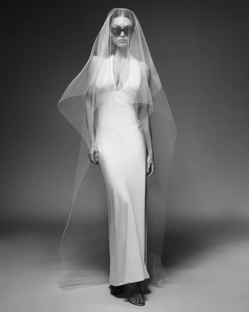 Monochrome portrait of a woman in a full-length white dress, sunglasses, and sheer veil, posing in a photo studio with a neutral backdrop.