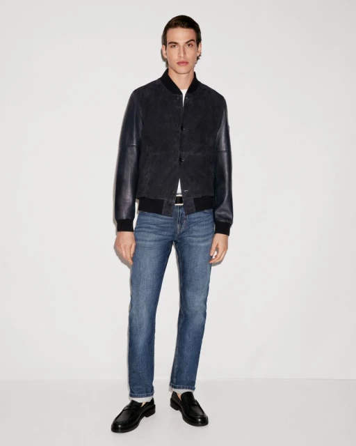 Full-length shot of an adult male model in a dark bomber jacket, blue jeans, and black loafers against a white backdrop in a studio.