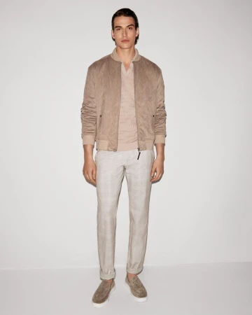 Full-length shot of an adult male model in a tan bomber jacket, beige collared shirt, and pants, standing against a white backdrop.