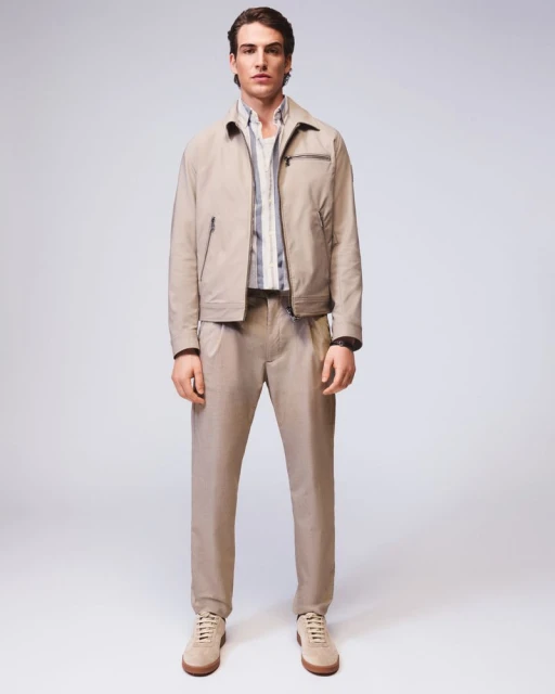 Full-length shot of an adult male fashion model in a tan jacket, vertically striped shirt, trousers, and suede shoes in a bright studio.