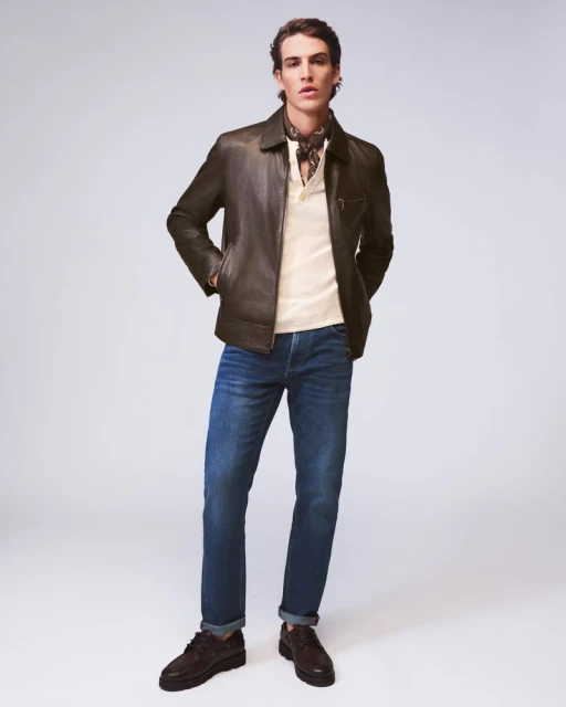 Man in a dark leather jacket, light-colored shirt, blue jeans, and dark shoes stands in a full shot against a neutral backdrop.