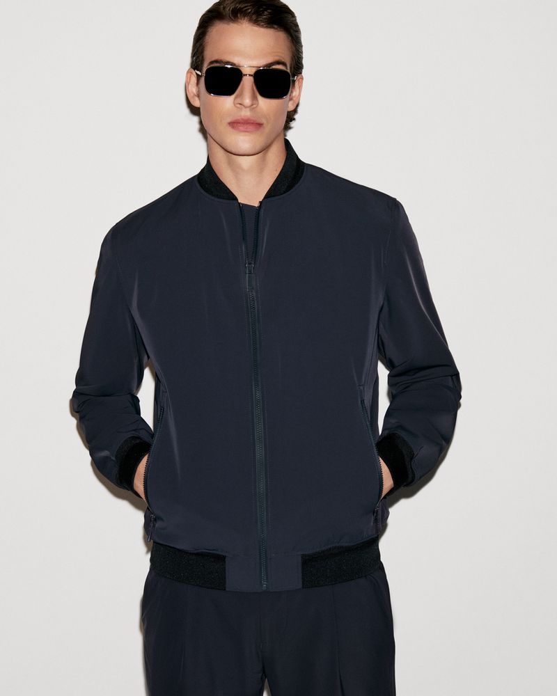 An adult male model poses in a navy blue bomber jacket and matching pants, accessorized with black sunglasses, against a neutral backdrop.