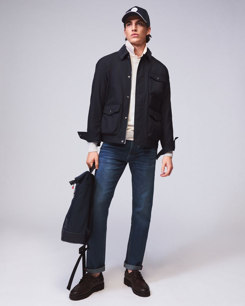 Male model wearing a black jacket, blue jeans, and baseball cap holding a backpack against a seamless grey backdrop.