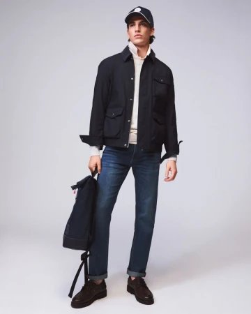 Male model wearing a black jacket, blue jeans, and baseball cap holding a backpack against a seamless grey backdrop.