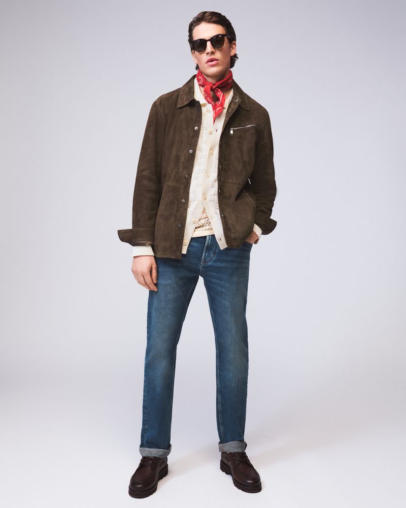 A stylish adult man in a brown jacket, blue jeans, and sunglasses stands in a photo studio with a red bandana, brown boots, and a white background.