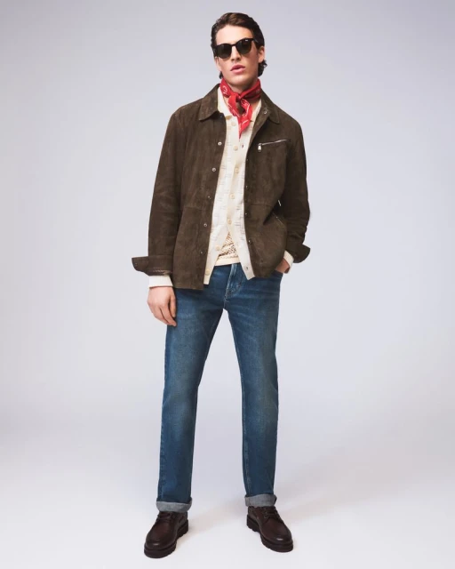 A stylish adult man in a brown jacket, blue jeans, and sunglasses stands in a photo studio with a red bandana, brown boots, and a white background.