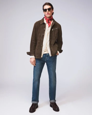 A stylish adult man in a brown jacket, blue jeans, and sunglasses stands in a photo studio with a red bandana, brown boots, and a white background.