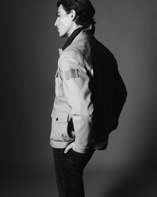 Monochrome studio portrait of a man in a jacket, turned to the side, with his hand in his pocket, against a dark background.