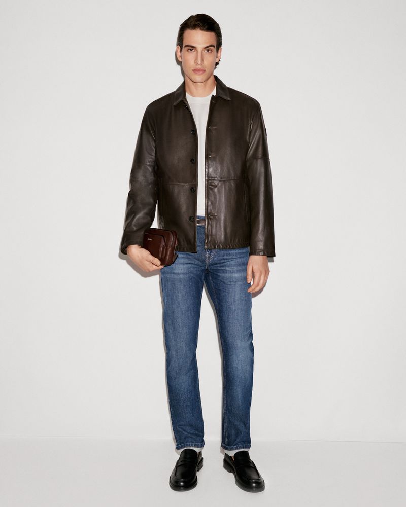 Full-length studio shot of a man in a dark leather jacket, jeans, and loafers, holding a small leather bag, against a white background.