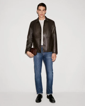 Full-length studio shot of a man in a dark leather jacket, jeans, and loafers, holding a small leather bag, against a white background.