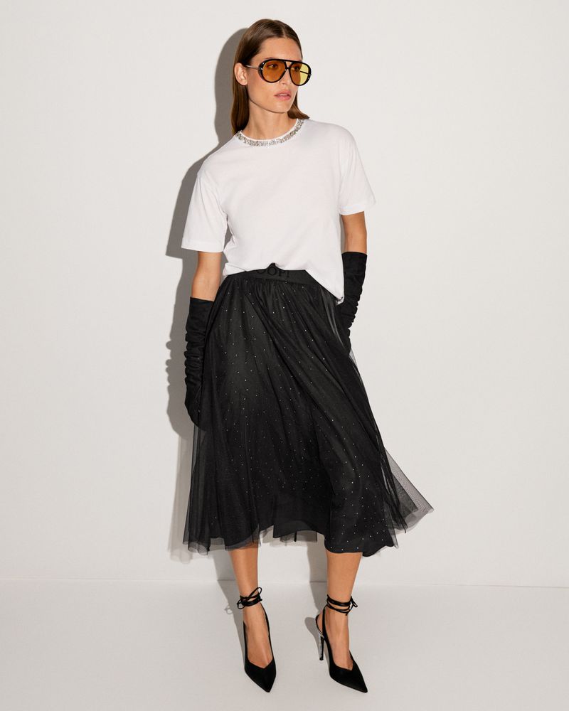 Woman in a white t-shirt and black tulle skirt poses in a studio. She wears gloves, sunglasses, and black high-heeled shoes with ankle ties.
