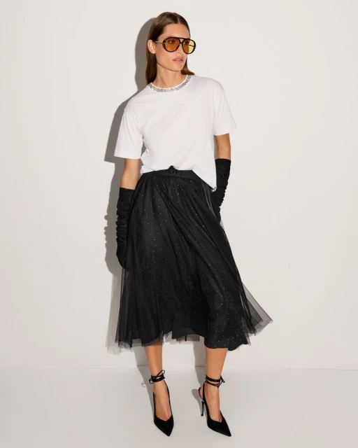Woman in a white t-shirt and black tulle skirt poses in a studio. She wears gloves, sunglasses, and black high-heeled shoes with ankle ties.
