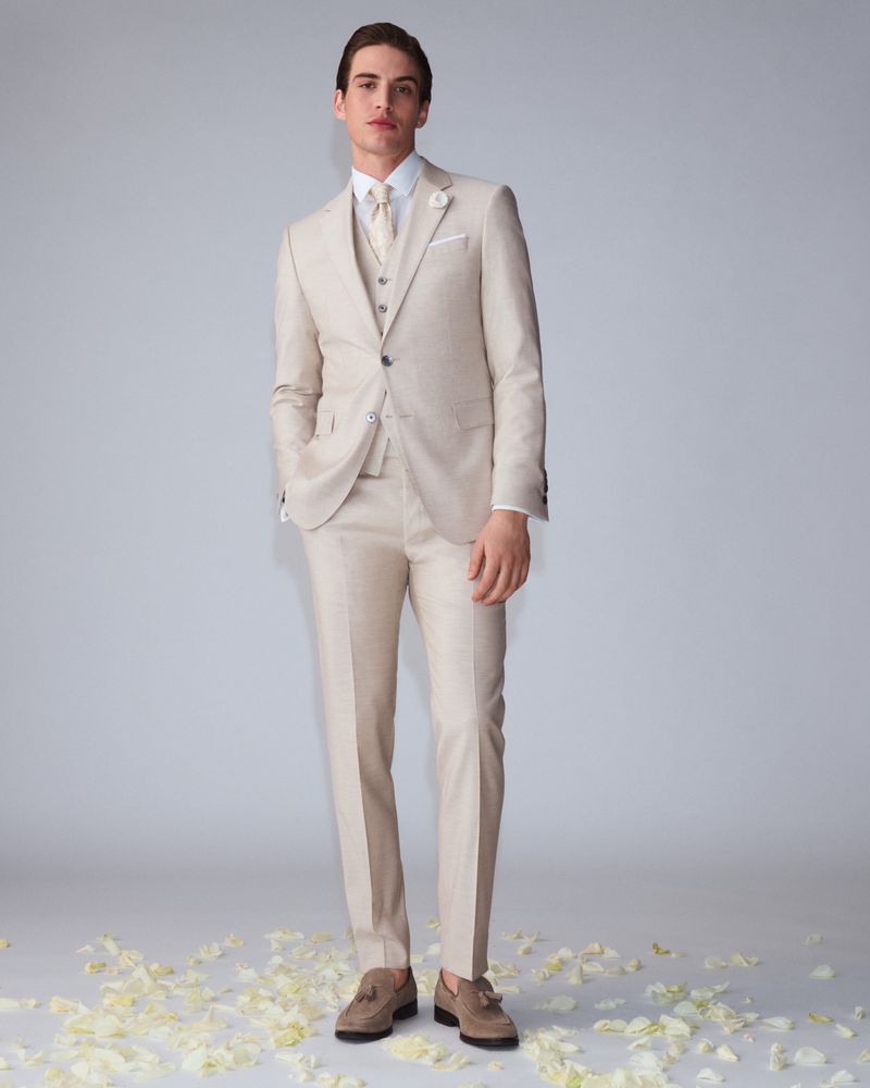 Full-length shot of a young adult male model in a tan three-piece suit, white shirt, and tan loafers, posing against a grey background.