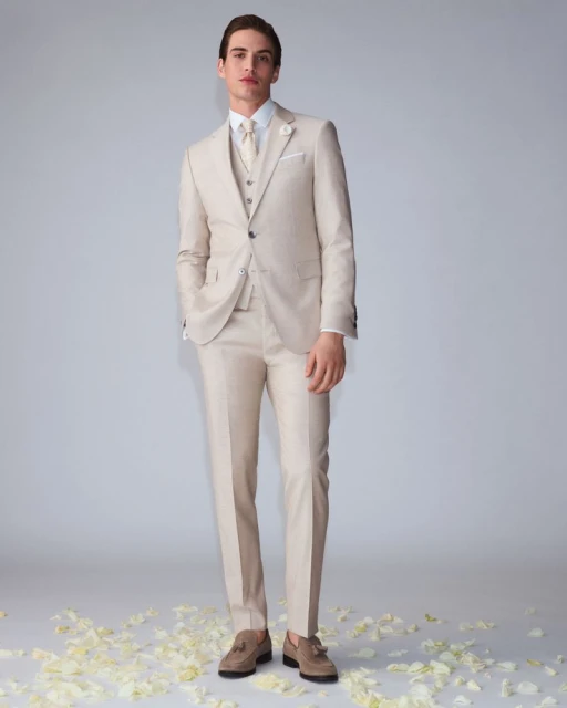 Full-length shot of a young adult male model in a tan three-piece suit, white shirt, and tan loafers, posing against a grey background.