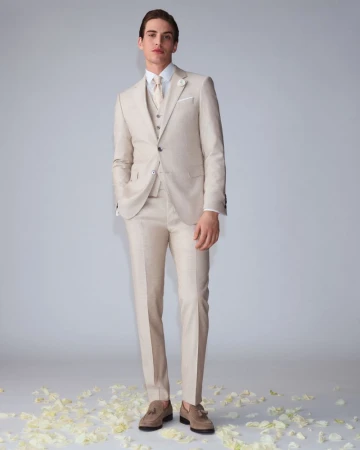 Full-length shot of a young adult male model in a tan three-piece suit, white shirt, and tan loafers, posing against a grey background.