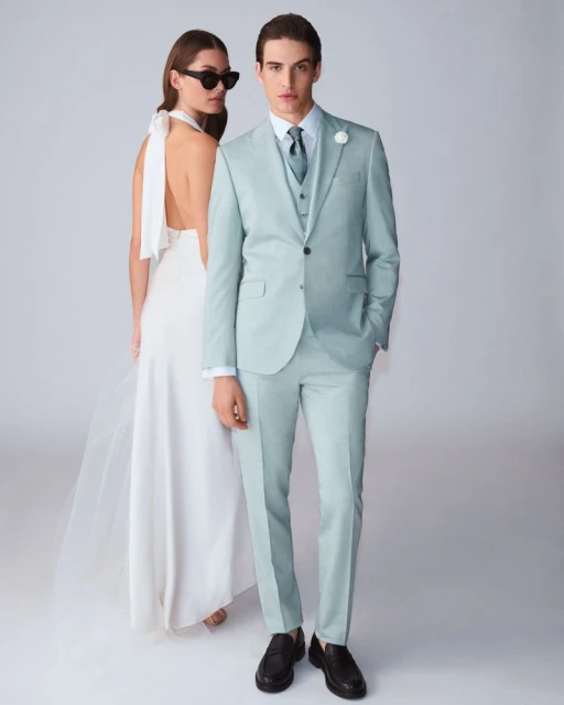 A man in a mint green suit and a woman in a white dress pose together in front of a white background in a fashion shot.