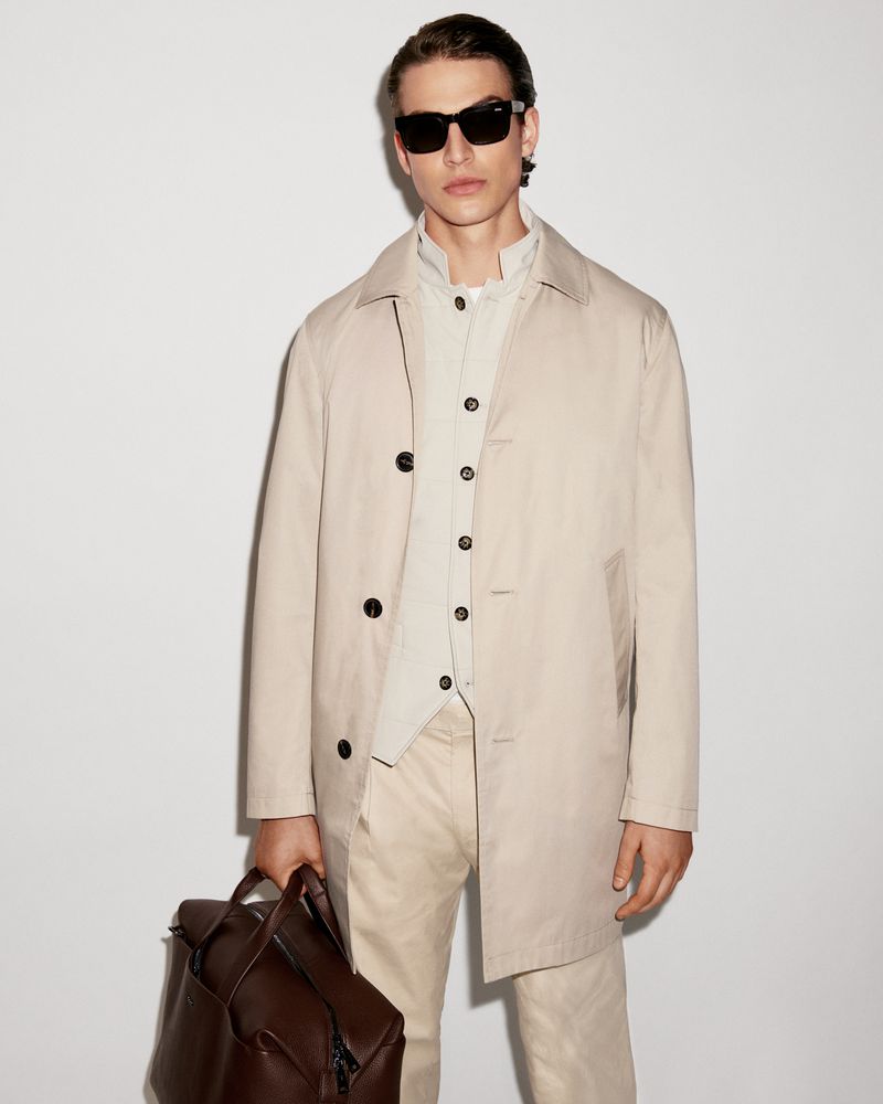 Man in a beige trench coat and sunglasses holds a brown leather backpack against a plain background, showcasing a modern, stylish look.