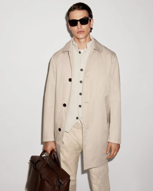 Man in a beige trench coat and sunglasses holds a brown leather backpack against a plain background, showcasing a modern, stylish look.