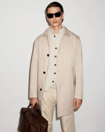 Man in a beige trench coat and sunglasses holds a brown leather backpack against a plain background, showcasing a modern, stylish look.