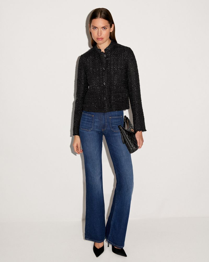 A woman in a black jacket and blue jeans stands in front of a white background, holding a black purse and wearing black heels.