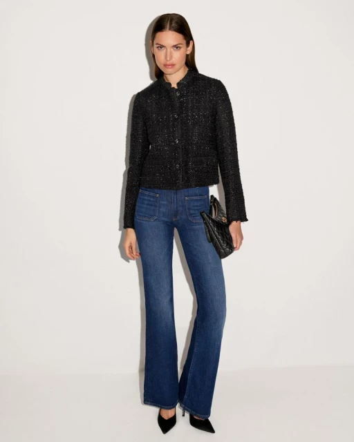 A woman in a black jacket and blue jeans stands in front of a white background, holding a black purse and wearing black heels.