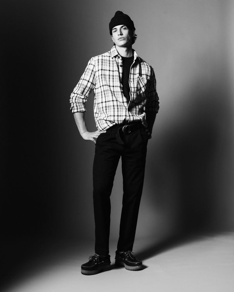 A model poses in black pants, a plaid shirt, and a beanie against a muted gradient backdrop in a studio fashion shot.