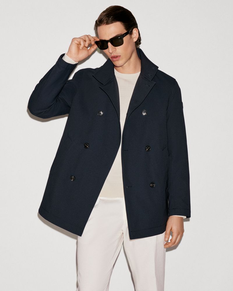 Man in navy peacoat and off-white pants poses indoors with sunglasses. Fashion editorial shot with neutral backdrop.