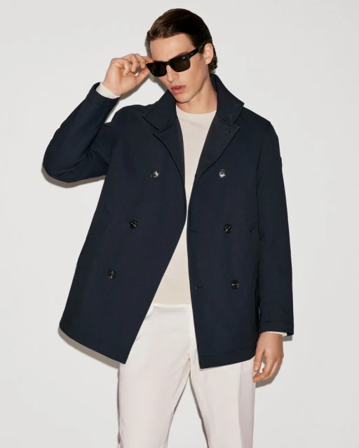 Man in navy peacoat and off-white pants poses indoors with sunglasses. Fashion editorial shot with neutral backdrop.