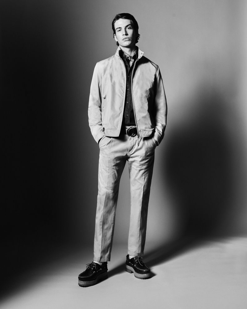 A full-length shot in black and white of a stylish male model in a jacket and trousers, posing confidently in a photo studio.