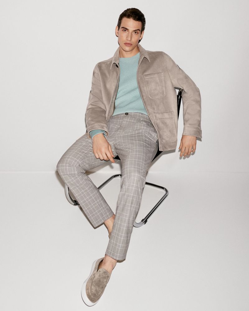 A stylish adult male wearing a tan jacket, light blue sweater, and patterned grey pants sits on a black chair in a bright studio.