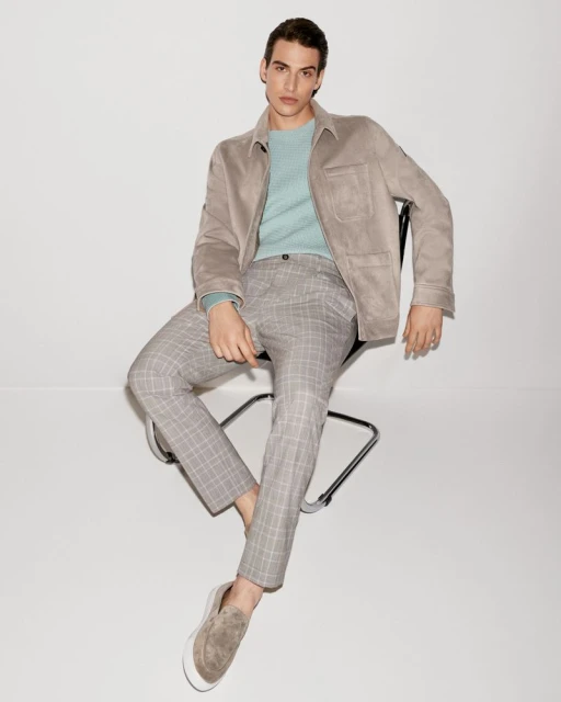 A stylish adult male wearing a tan jacket, light blue sweater, and patterned grey pants sits on a black chair in a bright studio.