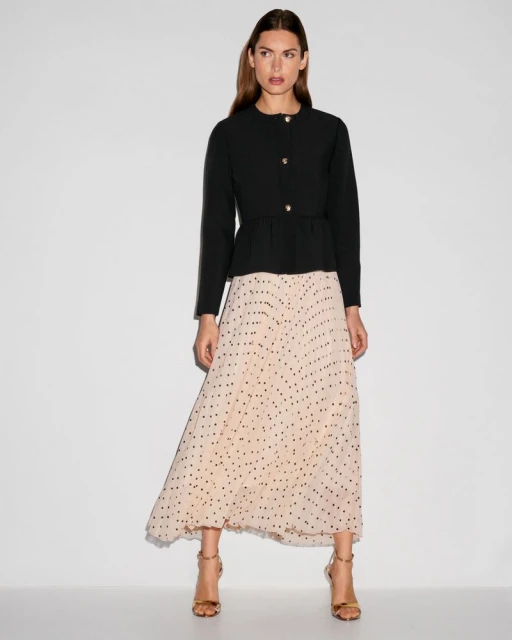 Full-length studio shot of a woman wearing a black peplum jacket with gold buttons and a long beige skirt with black polka dots.