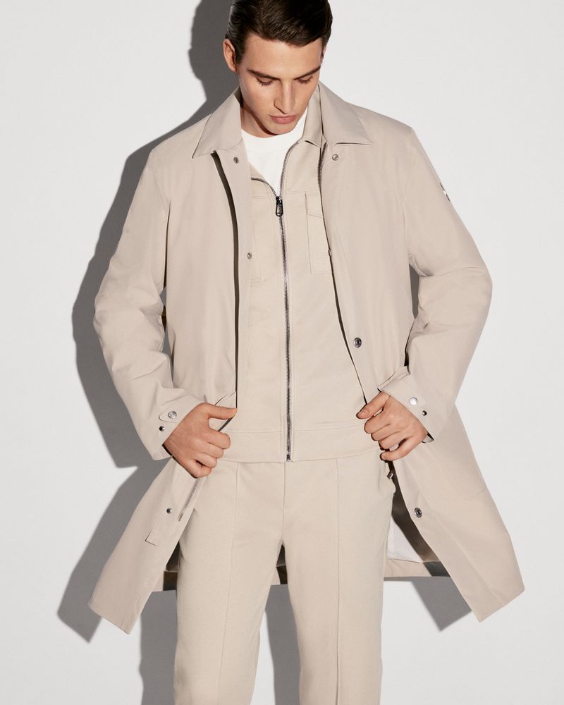 Man in a beige overcoat and matching suit posing in a bright photo studio. He has his hands in his pockets and looks down slightly.