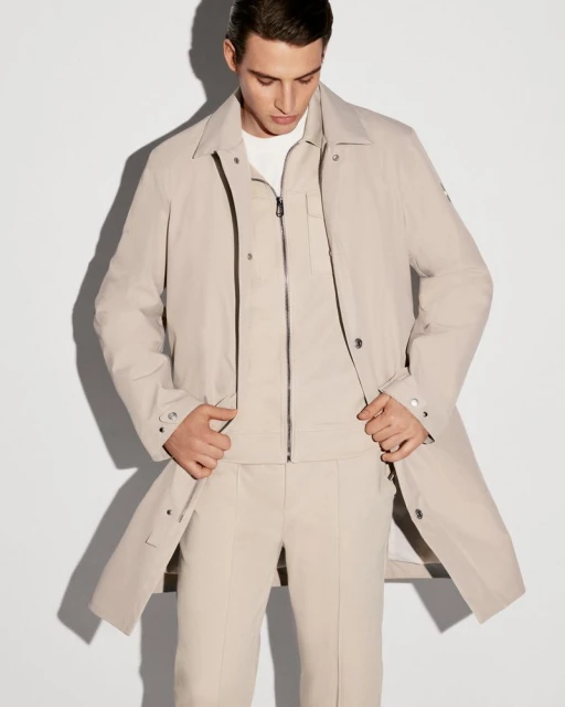 Man in a beige overcoat and matching suit posing in a bright photo studio. He has his hands in his pockets and looks down slightly.