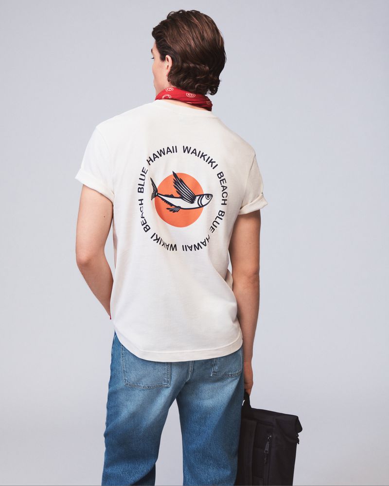 Back view of a man wearing a white t-shirt with a graphic print, blue jeans, and a red bandana, holding a black bag.