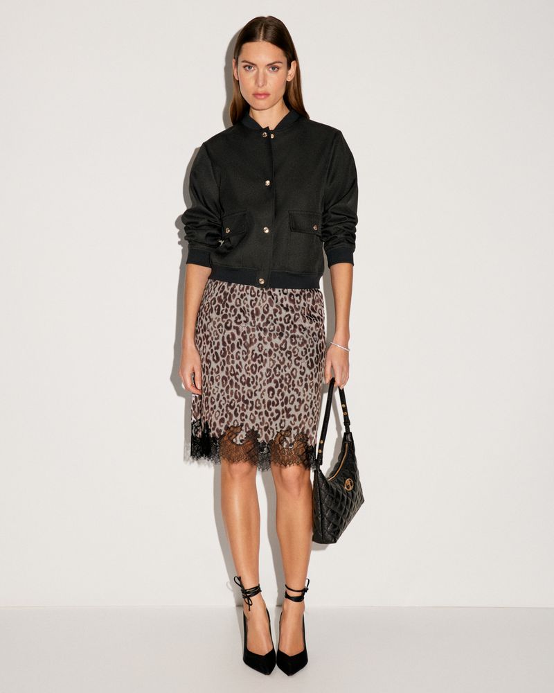 Fashionable woman in black bomber jacket, leopard print skirt, and high heels poses against a white backdrop, holding a black purse.
