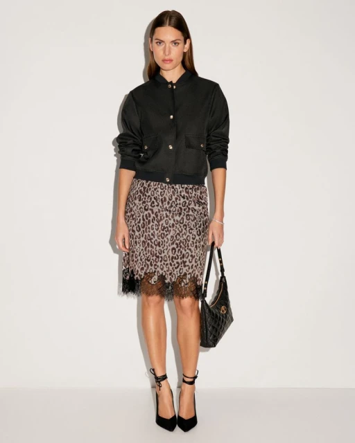 Fashionable woman in black bomber jacket, leopard print skirt, and high heels poses against a white backdrop, holding a black purse.