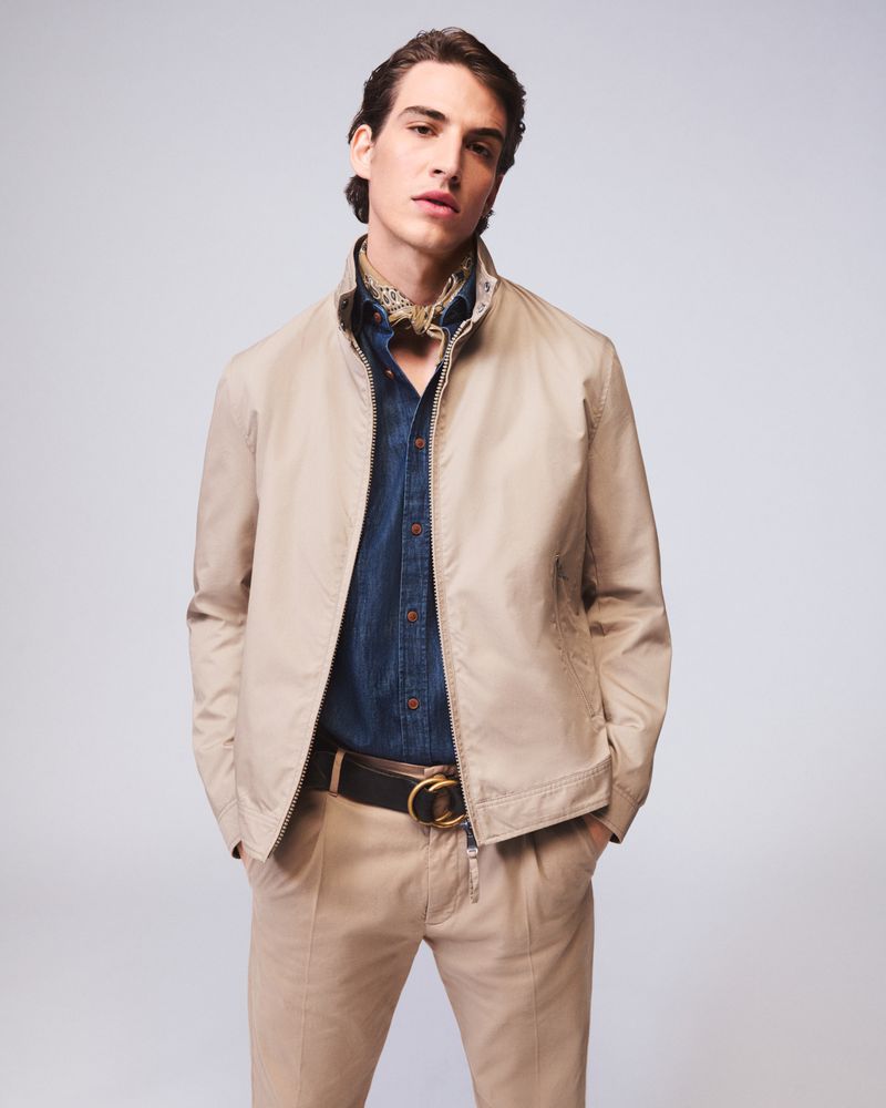 A light-skinned man in a jacket, button-down shirt, and tan pants poses against a neutral backdrop in a fashion shot.