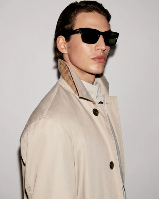 A man in a light brown coat and black sunglasses poses against a white wall. Head and shoulders shot, side view of a young adult.
