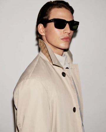 A man in a light brown coat and black sunglasses poses against a white wall. Head and shoulders shot, side view of a young adult.