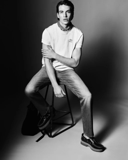 A young adult male sits on a metal stool in a studio wearing a white t-shirt, jeans, and black boat shoes in a medium shot.