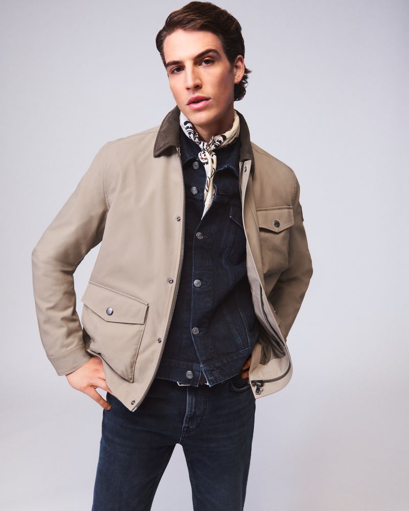A man poses in a tan jacket, blue denim shirt, and dark jeans in a studio setting, showcasing fashion apparel with a neutral background.