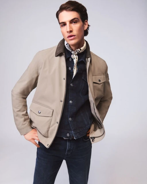 A man poses in a tan jacket, blue denim shirt, and dark jeans in a studio setting, showcasing fashion apparel with a neutral background.