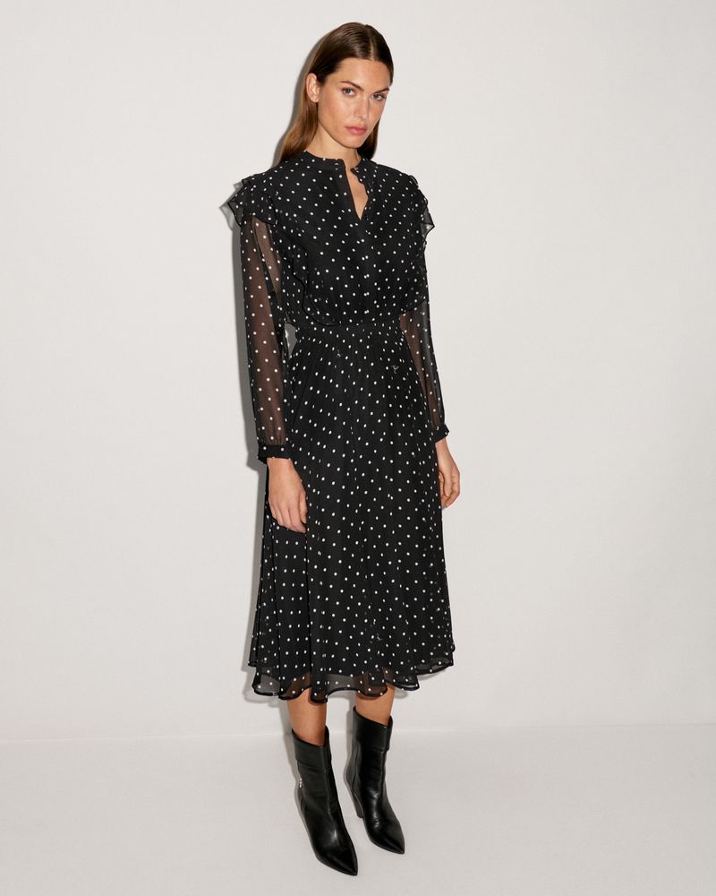 Elegant adult woman in a black polka dot dress and black heeled boots posing in a full shot against a white background.