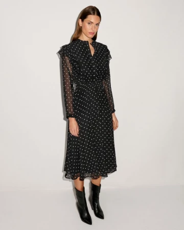Elegant adult woman in a black polka dot dress and black heeled boots posing in a full shot against a white background.