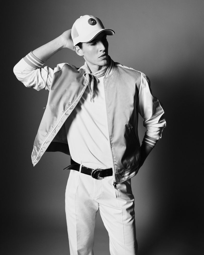 Black and white portrait of a young adult male in casual fashion, wearing a baseball cap, bomber jacket, and white pants, posing in studio.
