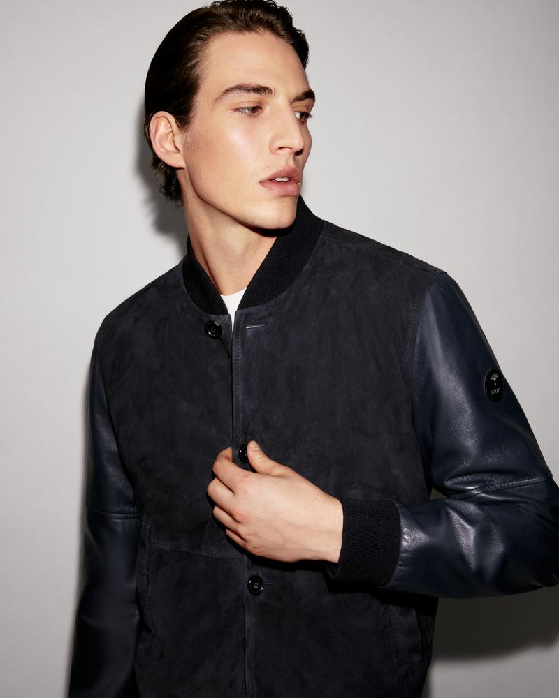 Portrait of a young adult male fashion model with dark hair and wearing a bomber jacket in a studio with neutral lighting.