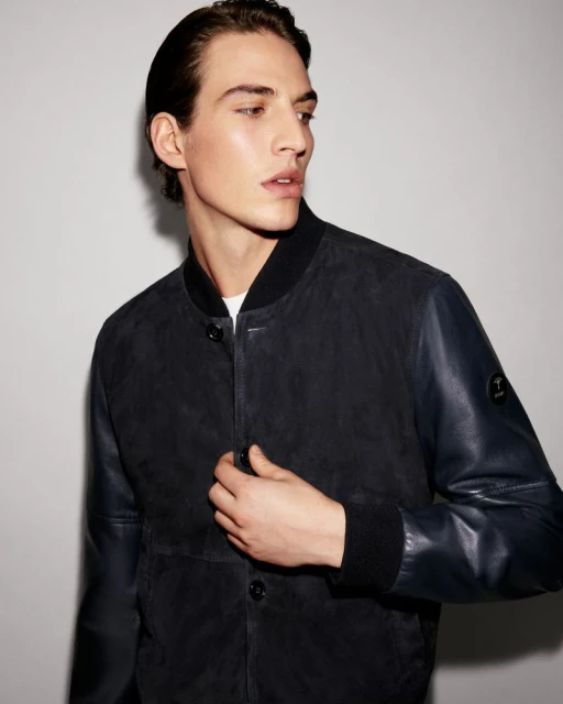 Portrait of a young adult male fashion model with dark hair and wearing a bomber jacket in a studio with neutral lighting.