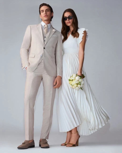 A stylish couple poses in coordinated formal wear against a neutral backdrop. The man wears a beige suit, while the woman wears a white dress and sunglasses.