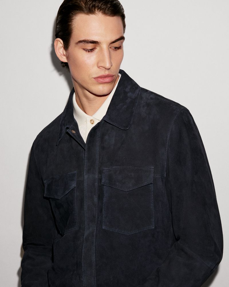 Portrait of a young adult male wearing a collared navy blue jacket with patch pockets against a white background in a studio setting.